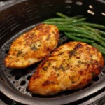 Two grilled chicken breasts in an air fryer basket with cooked green beans.