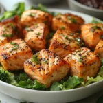 Grilled salmon bites garnished with herbs and black sesame seeds served over a bed of leafy greens.