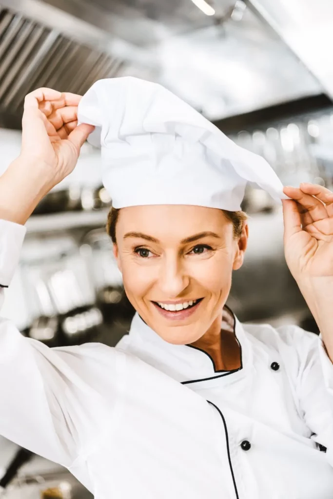 Smiling female chef adjusting her white hat in a professional kitchen.