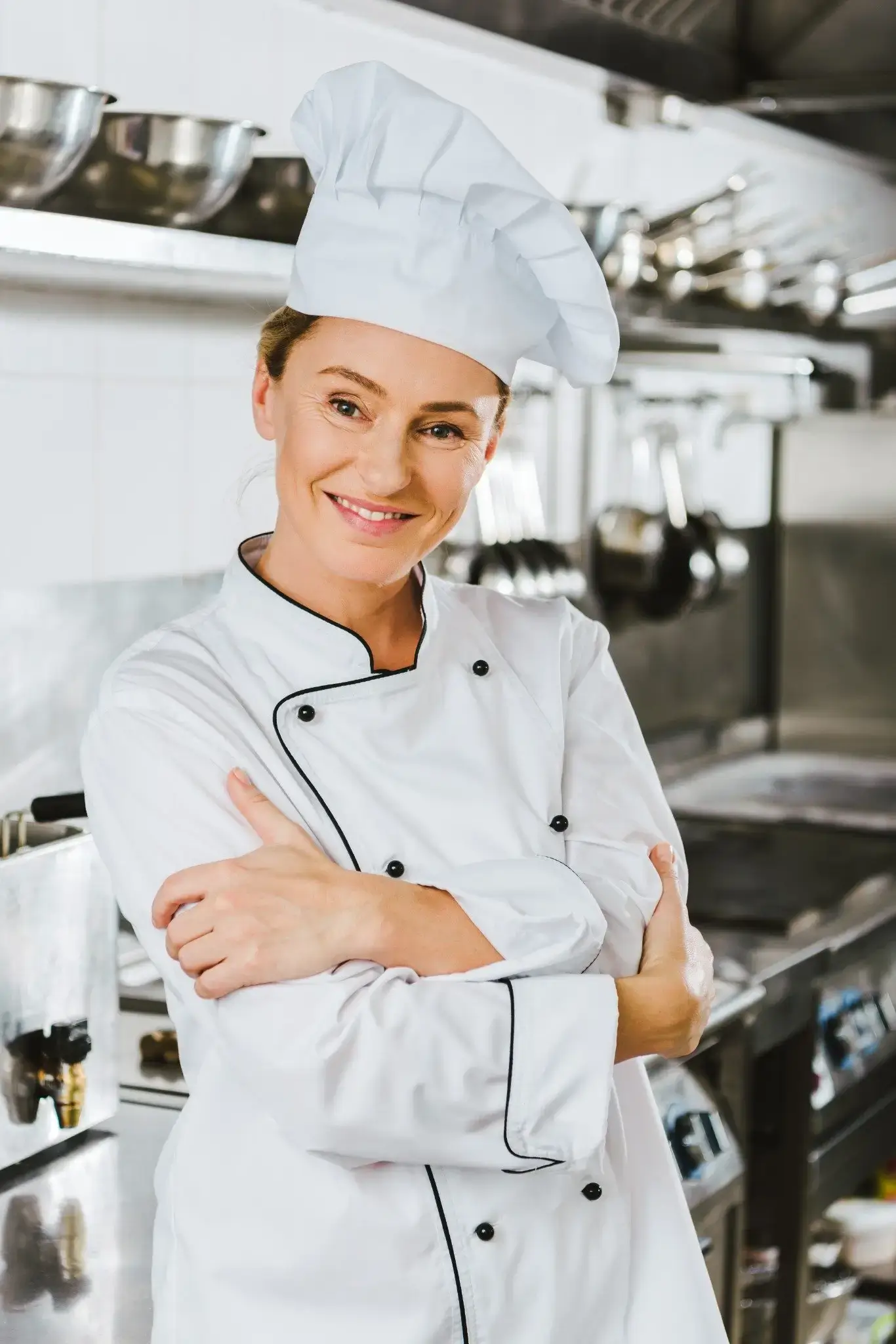 Confident female chef standing with arms crossed in a commercial kitchen.