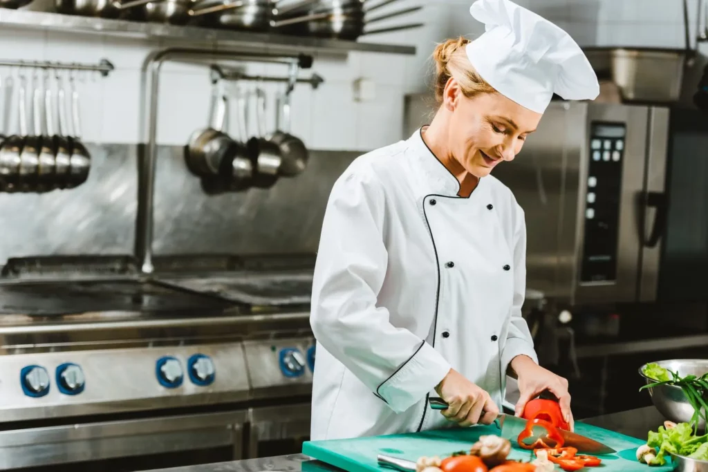 Female chef slicing a red bell pepper on a cutting board in a professional kitchen.