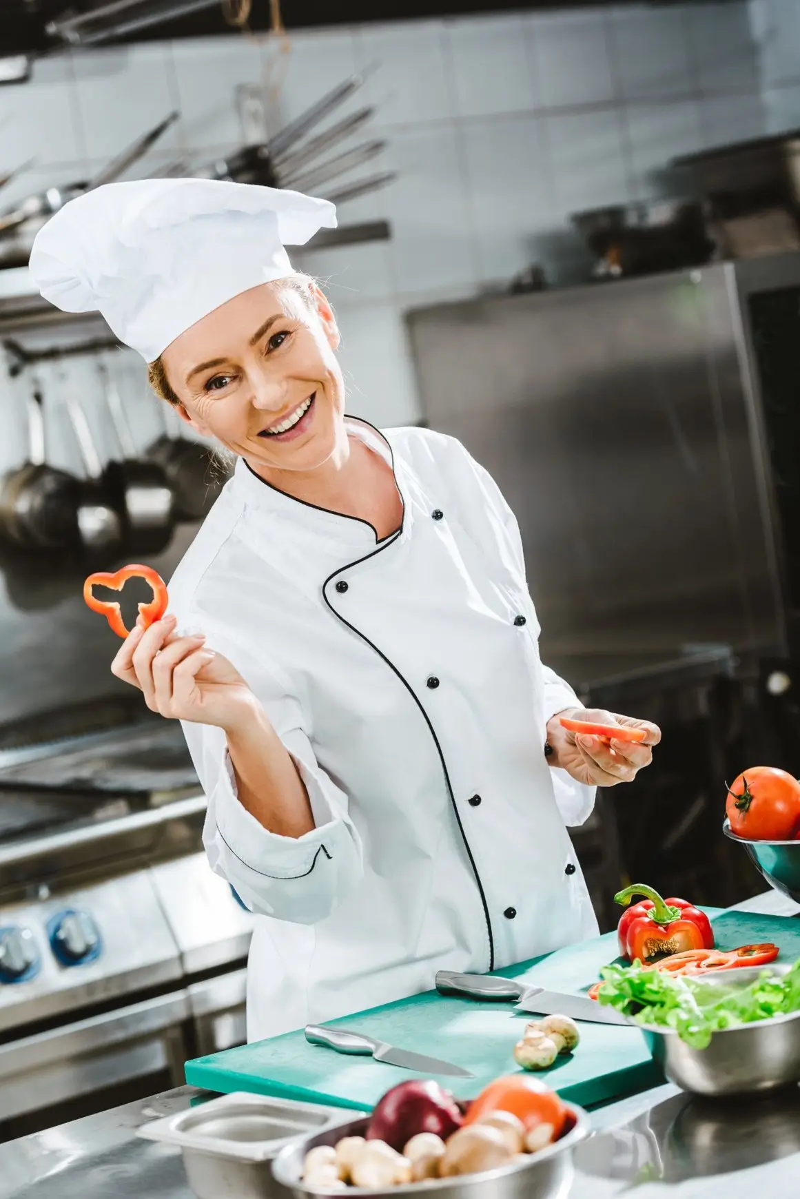 Smiling female chef in a professional kitchen holding a sliced red bell pepper.